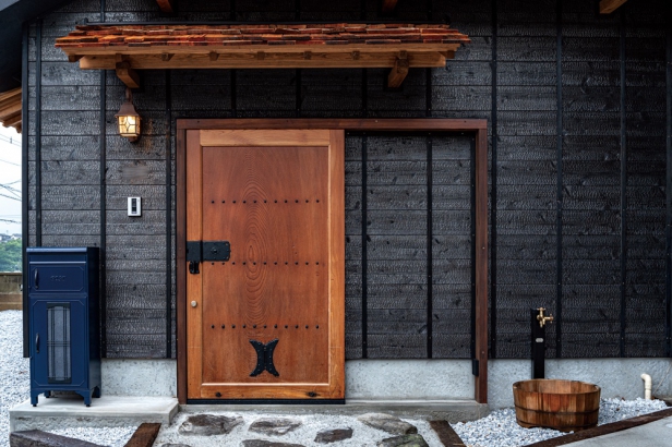 重厚な蔵戸や自然の色味を引き出した焼き杉の真っ黒な外壁が印象的な玄関。入口に用いた蔵戸は、張り合わせなどではなく、欅（ケヤキ）の輪切り一枚板を装飾したもの